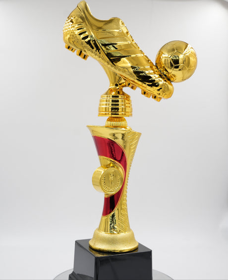 Full view of a shiny gold football trophy featuring a golden boot kicking a soccer ball, sitting on a red and gold stem with a black base.